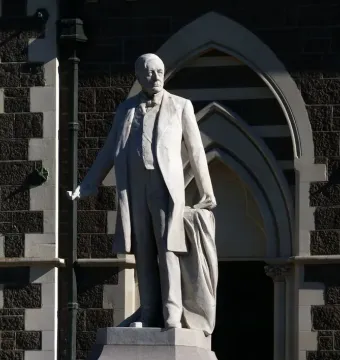 Rolleston Statue of a formally dressed man holding draped fabric beside a dark stone building.