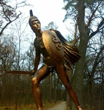 Bronze Greek warrior monument showing a helmeted soldier with a shield and raised sword.