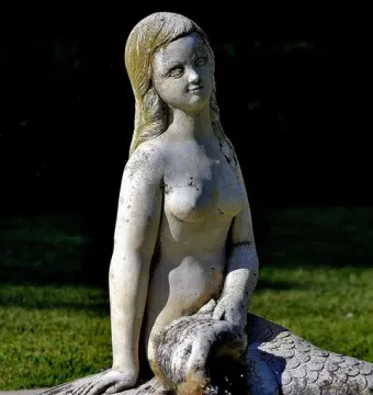 Weathered stone mermaid fountain statue with water flowing from the figure\'s hands.