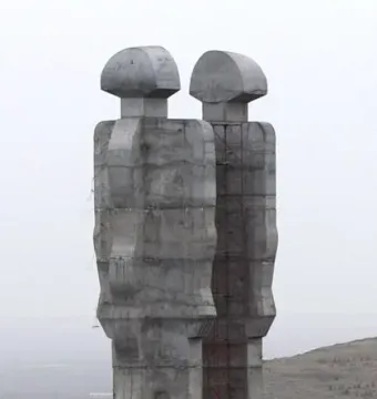 Two colossal abstract concrete figures of the Monument to Humanity stand on a hillside with scaffolding between them and two people at the base.