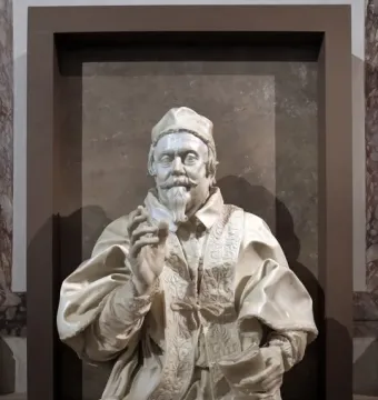 White marble bust of Pope Clement X with a raised right hand and a small book in his left hand.