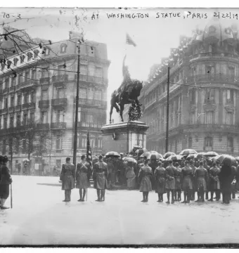 Crowd with umbrellas gathered before the Washington statue in a rainy square lined with ornate buildings.