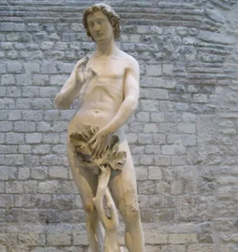 Adam, a slender marble statue with curly hair and foliage covering the groin, standing against a stone wall.