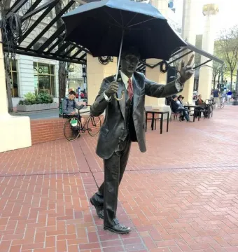 Allow Me, a dark bronze sculpture of a man in a suit holding a large umbrella and raising one hand.