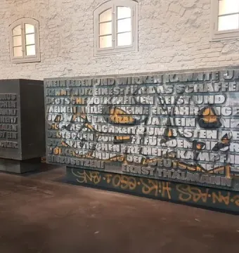 Two inscribed plaques from the Ernst Thälmann Memorial, one marked with orange graffiti, against a brick wall with arched windows.