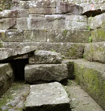Weathered stone blocks and a slanted slab inside an Etruscan tomb ruin, with patches of green moss.