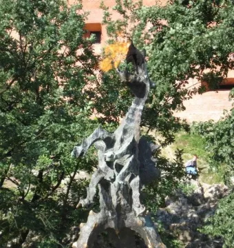 Wawel Dragon sculpture with a flame coming from its open mouth, surrounded by greenery and a brick building in the background.