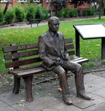 Alan Turing Memorial bronze statue of a man sitting on a park bench holding an apple, with an information board nearby.