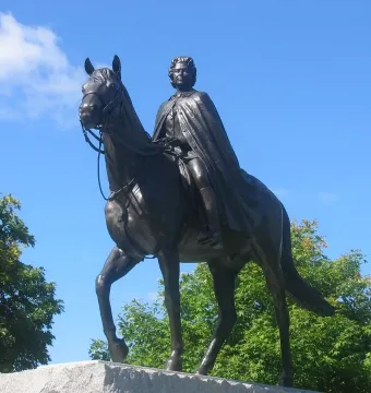 Elizabeth II equestrian statue on a stone pedestal with green foliage and blue sky behind it