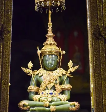 The Emerald Buddha seated on a golden pedestal beneath an ornate canopy with gilded columns and jeweled decorations.