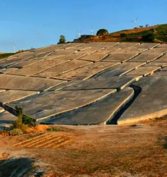 Il Grande Cretto’s cracked concrete slabs spread across a hillside beneath wind turbines and a blue sky.