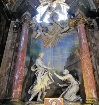 Noli Me Tangere sculpture showing a robed figure with an upraised hand facing a kneeling figure reaching out, set before a painted backdrop and framed by ornate columns.