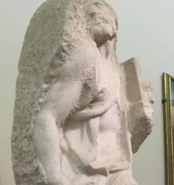 Unfinished marble sculpture of Saint Matthew emerging from a rough stone block, with detailed head, arm, and draped torso.