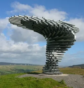 The Singing Ringing Tree sculpture made of stacked metal pipes stands on a grassy hill with rolling countryside and wind turbines in the distance.