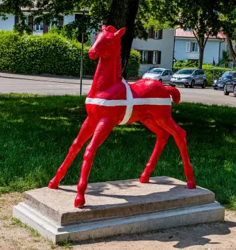 Holbeinpferd sculpture painted bright red with a white cross, standing on a concrete base in a grassy area.