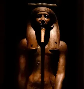 Wooden statue of King Hor with a pharaonic headdress and false beard on a black background.