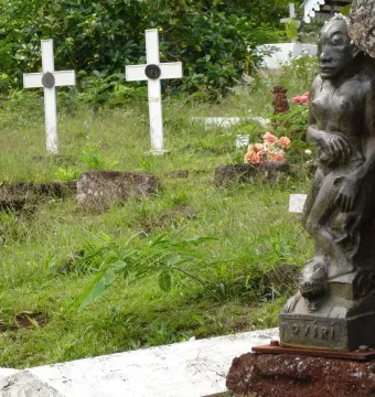 Oviri sculpture, a dark figure holding a smaller creature, standing in a grassy cemetery.