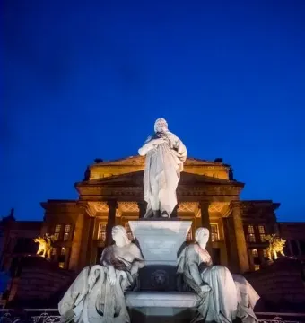 Schiller Monument illuminated at night in front of the Konzerthaus in Berlin, with seated allegorical figures beside the central statue.