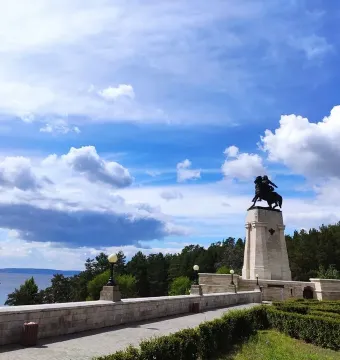 Tatishchev Monument on a tall stone pedestal beside a riverside path and low wall, with trees and water in the background.