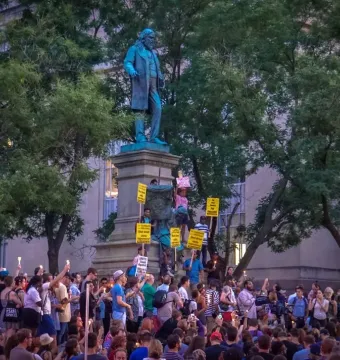 Albert Pike Memorial statue surrounded by a crowd holding candles and protest signs.