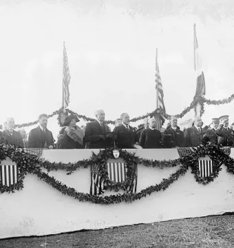 Dignitaries gathered on an outdoor platform decorated with garlands and shield emblems, with flags behind them and no statue visible.