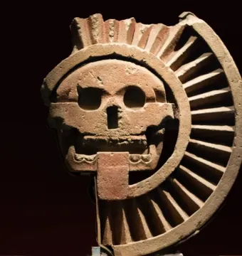 Disk of Mictlāntēcutli showing a skull-like face carved in reddish-brown stone with a spiky crown and ribbed fan-like elements.