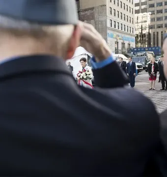 People gathered near the Katyń Memorial in an urban plaza, with one person saluting in the foreground and a woman holding white flowers in the distance.
