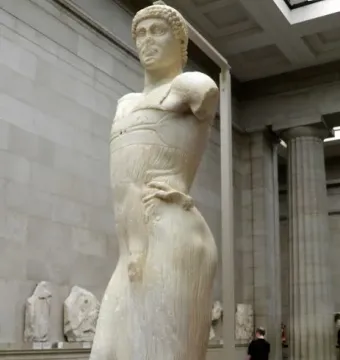 The Motya Charioteer standing on a red platform in a museum hall with columns and other artifacts nearby.