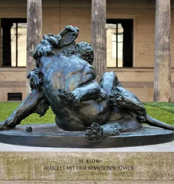 Bronze sculpture of Hercules grappling with the Nemean Lion on a stone pedestal outdoors.