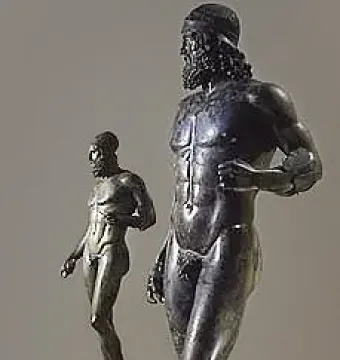 The Riace Bronzes displayed as two dark bronze male statues on light pedestals, one bearded figure in front and a smaller figure behind.