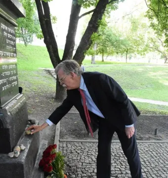 A man in a suit bends down to place a small stone at The Pit memorial, with red flowers and other stones at the base.