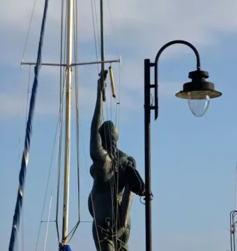 Verity sculpture seen from behind against a blue sky, framed by sailboat rigging and a lamppost.