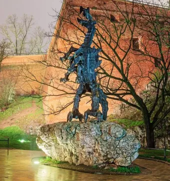 The Wawel Dragon sculpture stands on a large rock base beside a warmly lit brick wall and bare trees, with wet ground in the foreground.