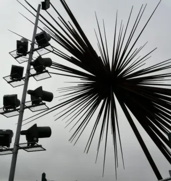 B of the Bang sculpture silhouetted against a grey sky beside a tall floodlight pole.