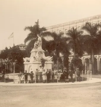 The Fuente de la India fountain in Havana with a seated female statue, adults and children gathered around it, and a large building and palm trees in the background.