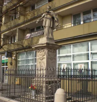 Stone statue of John of Nepomuk on an ornate pedestal behind a black spiked fence.