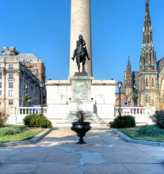 Lafayette Monument with a dark equestrian statue on a stone pedestal, a tall cylindrical monument behind it, and churches and buildings around the plaza.