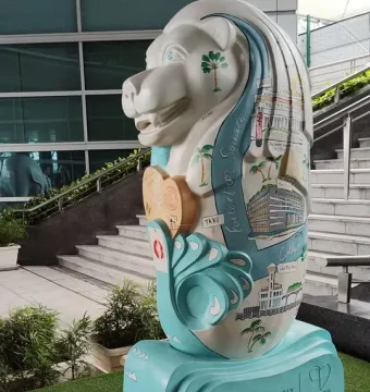 A painted Merlion sculpture with Singapore landmarks and palm trees, standing on a wave-shaped base near a staircase.