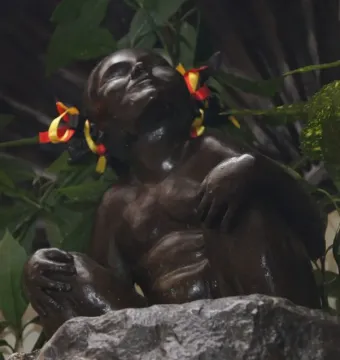 Jeanneke Pis bronze statue of a squatting young girl with ribbons in her hair, surrounded by green leaves and flowers.
