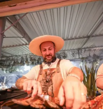 A bearded man wearing a straw hat and patterned bandana handles grilled food with tongs beside a Laçador graphic on his leather apron.