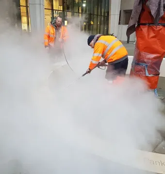 Workers clean the Pankhurst Memorial pavement with a high-pressure washer beside a wrapped vertical structure.