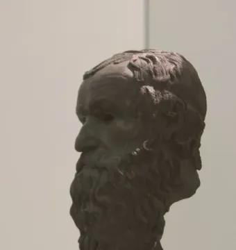 Dark patinated bronze head of the Porticello Philosopher in profile, with curly beard and hair on a light pedestal.
