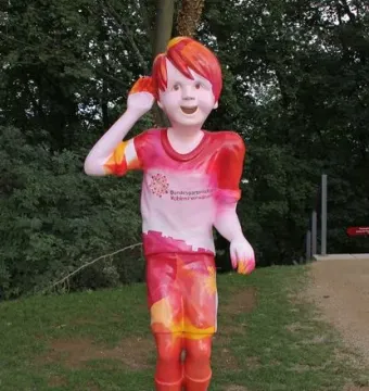 Schängel statue of a smiling boy with one hand raised to his ear, painted in red, pink, and yellow patterns.