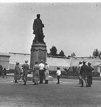 William Henry Seward statue on a tiered pedestal in Volunteer Park, with people gathered nearby and glass greenhouses in the background.