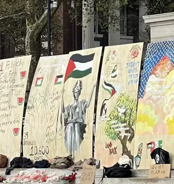 Lower portion of Alma Mater is partially visible behind protest signs and a metal barrier, with the base and part of the name visible.
