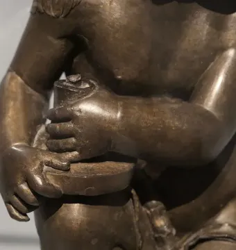 Close-up of a bronze Candelabra Angels figure with bare torso and arms, hands clasping a cylindrical object on a shallow disc.