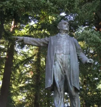 Harvey W. Scott statue of a man in a long coat pointing forward, set on a light-colored base among pine trees.