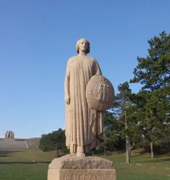 The La France 1918 stone sculpture stands on a pedestal under a clear blue sky, with a round shield in one hand and another stone figure in the distance.