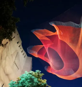 Janet Echelman’s 1.26 glows as a red and blue mesh sculpture suspended above trees at night.
