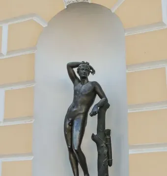 Apollino bronze nude male statue with one arm raised, standing in an arched niche beside a light yellow facade.